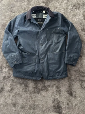 Blå vaxad rock från Levi's S - Snygg blå vaxad rock från Levi's i samarbete med Halley Stevensons. Rocken har tre framfickor, knäppning med tryckknappar och en krage i manchestertyg. Insidan är fodrad med mjuk, randig teddy för extra värme. Perfekt för kyliga dagar.