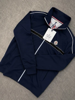 Moncler Ziptröja - Snygg marinblå tröja från Moncler med vit piping längs axlar och ärmar samt svart och vit rand över bröstet. Dragkedja framtill och två fickor. Insidan har ett coolt tryck med bergsmotiv och stor Moncler-logga. Perfekt för dig som gillar streetwear och exklusiva detaljer.