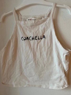 Vit croppad linnetopp Coachella - Säljer en vit croppad linnetopp från H&M Coachella-kollektionen. Toppen har raka axelband och texten 'COACHELLA' tryckt framtill. Perfekt för sommarens festivaler eller varma dagar.