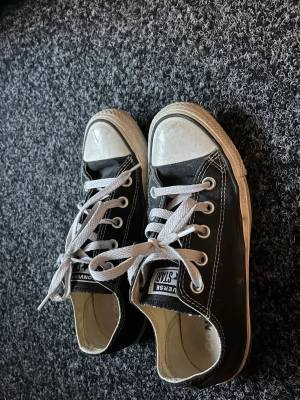 Svarta Converse All Star låga sneakers - Klassiska svarta Converse All Star sneakers med vit tåhätta och vita snören. Skorna har lågt skaft, vita sulor och tydliga kontrastsömmar. Perfekta för en avslappnad och tidlös stil. 