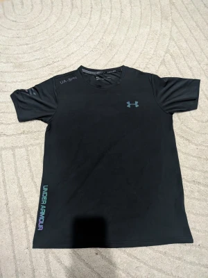Svart träningströja Under Armour - Svart kortärmad träningströja från Under Armour med rund hals och logga på bröstet. Tillverkad i ett lätt och snabbtorkande syntetmaterial, perfekt för intensiva träningspass. Diskret textdetalj på axeln och färgad logga på sidan.