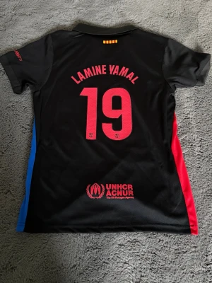 Svart FC Barcelona matchtröja Nike - Snygg svart FC Barcelona matchtröja med röda och blå detaljer på sidorna. Tryck med Lamine Yamal och nummer 19 på ryggen. Framtill finns Nike-logga, Barca-emblem och Spotify-logga. Tillverkad i lätt och ventilerande material, perfekt för fotbollsträning eller till vardags.