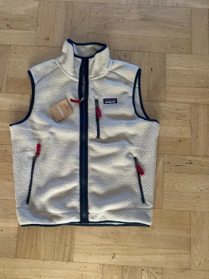 Patagonia väst - Säljer en vit fleeceväst från Patagonia med marinblå dragkedjor och detaljer samt röda zippar. Västen har hög krage, tre fickor med dragkedja och mjukt, fluffigt material. Perfekt för lager-på-lager och outdoor-vibe.