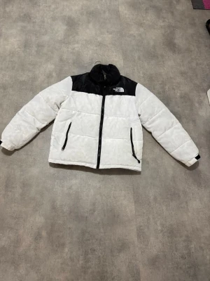 Vit och svart pufferjacka The North Face - Säljer en snygg vit pufferjacka från The North Face med svarta detaljer över axlar och krage. Jackan har dragkedja framtill, två sidofickor med dragkedja och hög krage. Perfekt för kalla vinterdagar och riktigt trendig stil. Har inte använt den än så länge eftersom har redan en som är svart. 