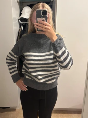 Gråvit randig stickad tröja - Mysig stickad tröja i grått och vitt med breda horisontella ränder över bröstet och ärmarna. Tröjan har en avslappnad passform, rund halsringning och ribbade muddar vid ärmslut och nederkant. Perfekt för kyliga dagar och enkel att matcha.