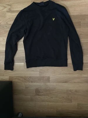 Svart sweatshirt med gul logga - En stilren svart sweatshirt med rund hals och lång ärm. På bröstet finns en liten gul broderad logga som detalj. Tröjan har ribbade muddar vid ärmslut och nederkant. Perfekt för en avslappnad och snygg look.