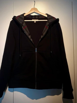 BB hoodie med rutigt foder - Säljer en svart hoodie med dragkedja och huva. Insidan av huvan och jackan har ett grått och rött rutigt foder som ger en snygg kontrast. Hoodien har fickor framtill och justerbara snören i huvan. Perfekt för dig som gillar en enkel men stilren look.