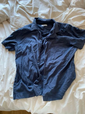 Mörkblå skjorta från These Glory Days - Säljer en mörkblå skjorta från These Glory Days i storlek S. Skjortan har korta ärmar, klassisk krage, bröstficka och knappar framtill. Perfekt till jeans eller shorts för en clean och enkel stil.