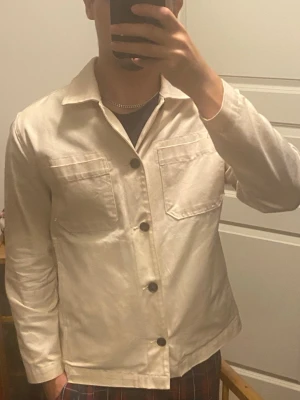 Beige overshirt från Mads nørgaard - Snygg beige overshirt från Mads nørgaard med fyra stora svarta knappar framtill och två bröstfickor. Rak passform och klassisk krage, perfekt att bära över en t-shirt. Materialet ser ut att vara bomull eller en bomullsblandning. Enkel och stilren design som passar till det mesta.