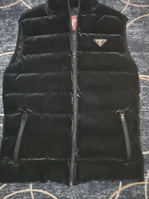 Svart dunväst från Prada - Stilren svart dunväst från Prada med quiltad design och hög krage. Västen har två dragkedjeförsedda fickor framtill och en diskret logotyp på bröstet. Perfekt för lager-på-lager och håller dig varm under kyliga dagar.