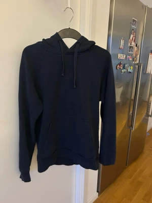 Mörkblå hoodie med huva - En enkel och stilren mörkblå hoodie med huva och dragsnören. Hoodien har en stor känguruficka framtill och långa ärmar. Perfekt för chill dagar eller när du vill ha en avslappnad look. Materialet känns mjukt och bekvämt mot huden.