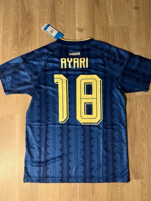 Blå Sverige fotbollströja  - Säljer en blå fotbollströja med Sveriges landslagsemblem och nummer 18 i gult. Tröjan har gul krage, tre gula ränder på axlarna och mönstrad design. Namnet 'Ayari' står på ryggen. Tillverkad i lätt och ventilerande material. HELT NY!