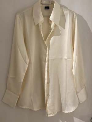Klassisk vit skjorta i silkeslook - Stilren skjorta med lång ärm och knappar framtill. Skjortan har en klassisk krage och är tillverkad i ett material med silkeslen känsla som ger en elegant look. Perfekt för dig som gillar tidlös och clean stil.