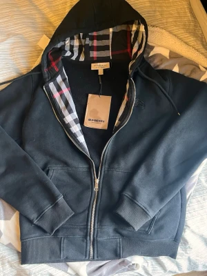 Svart zip-hoodie från Burberry - Snygg svart hoodie från Burberry med dragkedja och klassiskt rutigt foder i huvan. Tröjan har ribbade muddar, fickor framtill och diskret Burberry-logga broderad på bröstet. Perfekt för dig som gillar stilrena och exklusiva plagg.