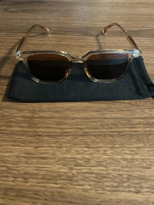Raybandliknande solglasögon  - Snygga solglasögon med transparent, fyrkantig båge och mörka glas. Bågarna har en lätt beige ton och glasen är svarta. Perfekta för soliga dagar och ger en cool vibe till din outfit. Medföljer en svart tygpåse för förvaring. Det ska vara uv skydd men har inte testat för uv skydd