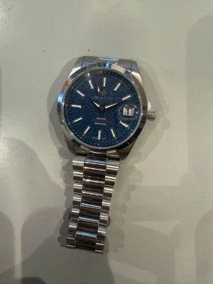 Stilren silverfärgad klocka med blå urtavla - Säljer en snygg klocka i silverfärgat metallarmband och blå urtavla. Klockan har tydliga indexmarkeringar, datumfönster och klassisk rund form. Perfekt accessoar för dig som vill ha en stilren look.