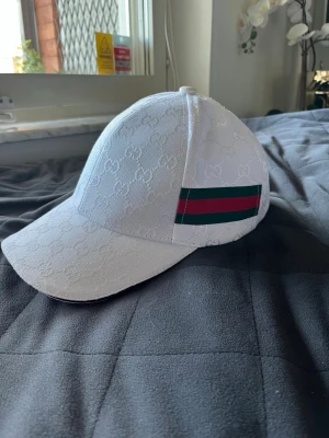 Vit Gucci keps med GG-mönster - Snygg vit keps från Gucci med det klassiska GG-mönstret i tyget och en ikonisk grön-röd rand på sidan. Justerbar rem bak i svart läder med Gucci-logga. Perfekt accessoar för att lyfta din outfit och ge en lyxig touch.