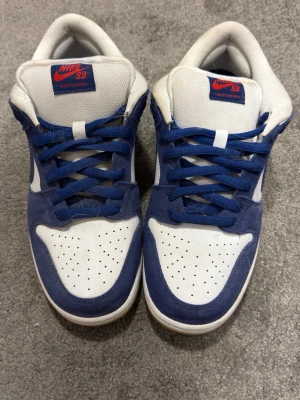 Nike SB Dunk Low blå/vit sneakers - Nike SB Dunk Low sneakers i blått och vitt med röd Nike-logga på plösen. Ovandelen är i mocka och textil, med perforerad tåbox för ventilation. Klassisk låg siluett och vita sulor. Perfekta för dig som gillar streetwear och skate. Från 🇨🇳