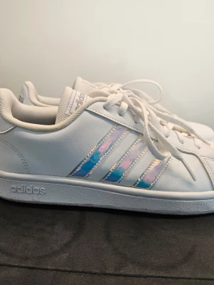 Adidas vita sneakers - FV8487  Vita sneakers med snörning. Skorna har tre ränder på sidorna i en skimrande, holografisk design som skiftar i blått och lila. Adidas-logotypen finns på plösen, hälen och yttersulan.  Endast använda ett fåtal gånger. Säljes då dem inte kommer användas mer.