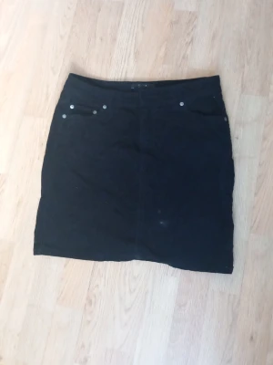 Svart manchesterkjol med fickor - Säljer en svart kortkjol i manchester med fickor både fram och bak. Kjolen har en enkel design med små silvriga nitar vid fickorna och är perfekt för dig som gillar en klassisk men trendig look.