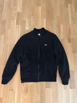 Svart Lacoste tröja med dragkedja  - Snygg svart ziptröja med hög krage och dragkedja hela vägen. Tröjan har en liten broderad logga på bröstet och ribbade muddar vid ärmslut och nederkant. Perfekt till chill eller streetwear-stil.