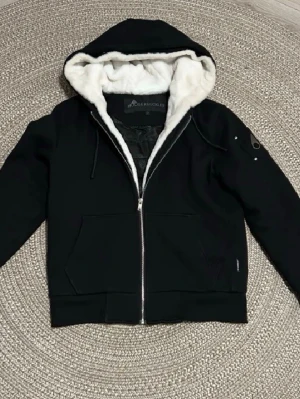 Svart hoodie med vit fuskpäls - Säljer en svart hoodie med dragkedja och vit fuskpäls i huvan. Hoodien har två stora fickor framtill, justerbar dragsko i huvan och ribbade muddar. Perfekt för dig som vill ha något både snyggt och mysigt till vardags. Materialet är mjukt och varmt.