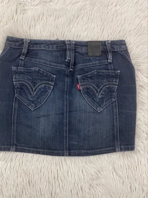Levi's Low-waist denim mini skirt W29 - Säljer en mörkblå jeanskjol i klassisk modell med två stora bakfickor och bälteshällor. Kjolen har en kort passform och är tillverkad i denim med slitningar för en cool look. Perfekt till sneakers eller boots.       Storleken är W29. Använd ett par gånger, men inget märkbart.
