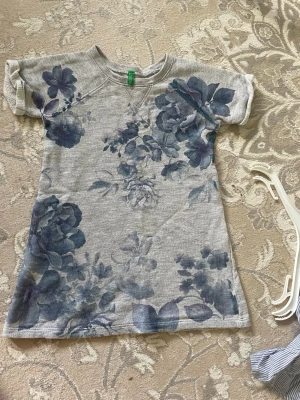 Grå blommig t-shirt med blå toner - Säljer en grå t-shirt med stora blå blommor på framsidan. T-shirten har korta ärmar och en klassisk rund halsringning. Materialet känns mjukt och skönt, troligen bomull. Perfekt för dig som gillar en avslappnad men ändå snygg stil.