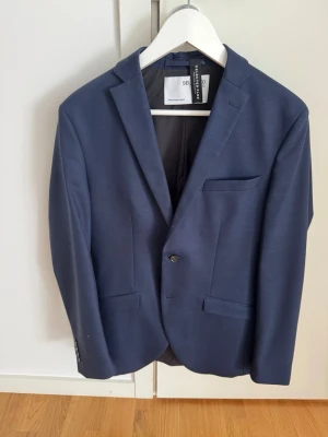 Mörkblå kavaj Selected Homme - Stilren mörkblå kavaj från Selected Homme med klassisk passform. Kavajen har två framfickor med lock, en bröstficka och enkelknäppning med två knappar. Materialet ser ut att vara en slät och lätt tygblandning, perfekt för en dressad look.