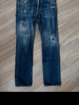 Blå slitna jeans från Dsquared2 - Säljer ett par blå jeans från Dsquared2 med distressed detaljer, hål och slitningar samt vita färgstänk. Jeansen har rak passform, klassiska fem fickor och knappgylf. Perfekta för en avslappnad och trendig look.