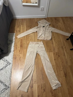 Beige juicy couture set - Mysigt loungewear-set i beige velour med matchande hoodie och byxor. Hoodien har dragkedja och broderad detalj framtill. Byxorna har resår i midjan och raka ben. Perfekt för chill dagar hemma eller när du vill vara extra bekväm.
