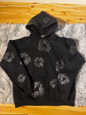 Svart hoodie från Denim Tears - Säljer en svart hoodie från Denim Tears med gråa blommönster över hela tröjan. Hoodien har en stor huva och en klassisk magficka framtill. Tillverkad i mjukt bomullsmaterial som är skönt att bära. Perfekt för dig som gillar streetwear och vill sticka ut.