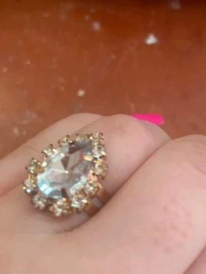 Guldig ring med stor droppformad sten - Snygg ring i guldfärg med en stor, droppformad klar sten i mitten. Runt stenen sitter mindre gnistrande stenar som ger extra bling. Perfekt för dig som gillar statement-smycken och vill sticka ut lite extra.