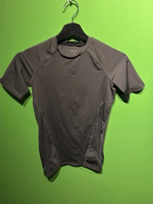 Base layer compresion shirt från Gymshark - Säljer en olivgrön, tight och kortärmad träningströja från Gymshark. Tröjan har rund halsringning och diskret logga på bröstet. Tillverkad i ett stretchigt funktionsmaterial som passar perfekt till gymmet eller löpning.