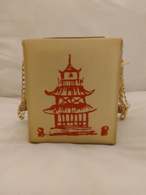 Guldfärgad väska i formen av take out kartong för kinamat. - Unik guld/ beige handväska i med röd pagodillustration på framsidan. Designen är i form av en klassisk take out kartong för kinamat. Väskan har guldfärgad kedjerem som kan användas kort, crossbody eller tas bort helt.Slät yta i konstläder. Perfekt för dig som vill sticka ut med en kreativ accessoar