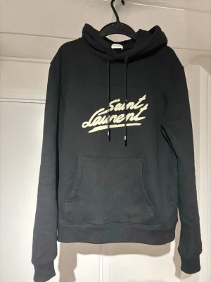 Svart Saint Laurent hoodie med tryck - Snygg svart hoodie från Saint Laurent med vit logga i graffiti-stil på bröstet. Klassisk känguruficka framtill och justerbar huva med snören. Perfekt för dig som gillar streetwear och vill ha en stilren men ändå statement look.