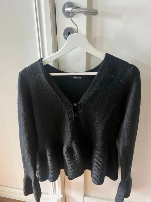 Svart kofta med volangkant - Snygg svart kofta med v-ringning och knäppning framtill. Koftan har långa ärmar och en unik volangkant nedtill som ger en trendig look. Tillverkad i mjukt stickat material som känns skönt mot huden.