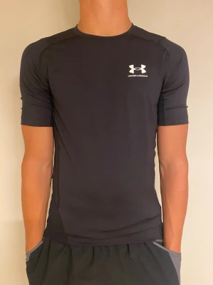 Svart kompressionströja Under Armour - Säljer en svart kompressionströja från Under Armour med HeatGear-teknologi. Tröjan har korta ärmar, rund hals och en tight passform som passar perfekt till träning. Logga i vitt på bröstet. Materialet är stretchigt och andas bra.