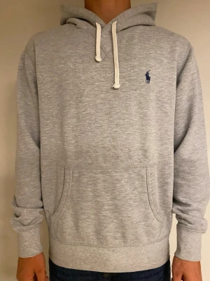 Grå hoodie från Polo Ralph Lauren - Säljer en stilren grå hoodie från Polo Ralph Lauren med klassisk huva och vita dragsnören. Tröjan har en känguruficka framtill och den ikoniska blå loggan broderad på bröstet. Tillverkad i mjukt bomullsmaterial som är skönt mot huden.