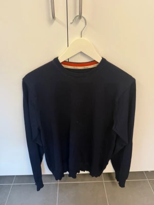 Stenströms Mörkblå rundhalsad tröja, stl M i 100% Merinoull  - En stilren mörkblå tröja med rund halsringning och diskret orange detalj på insidan av kragen. Tröjan har långa ärmar och ribbade muddar vid ärmslut och nederkant. Perfekt för lager på lager och enkel att matcha med olika outfits.