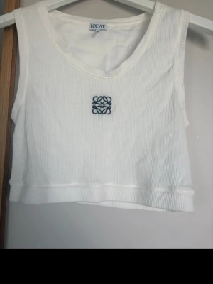 Loewe cropped anagram top  - Säljer vit ribbad linnetopp från Loewe med broderad logga framtill. Toppen är croppad och ärmlös, tillverkad i mjuk bomull med lite elastan för stretch. Perfekt för en trendig och avslappnad look.Toppen är använd en gång ståndet är som ny. Nypris är 3500kr. Kvitto från Net a porter finns om så önskas 