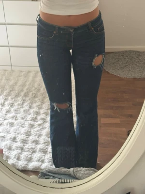 Mörkblå bootcut jeans med slitningar - Säljer ett par mörkblå bootcut jeans med slitna detaljer på låret och knät. Jeansen har låg midja och klassisk femficksdesign. Materialet är denim med stretch för skön passform. Perfekta för en avslappnad och trendig look.
