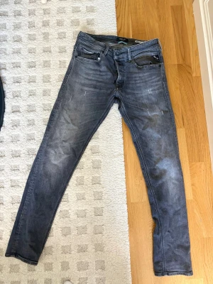 Grå slim fit jeans från Replay - Snygga grå slim fit jeans från Replay med slitningar och klassisk femficksdesign. Jeansen har en tvättad look och är tillverkade i bomull med lite stretch för extra komfort. Perfekta för en avslappnad och trendig stil.