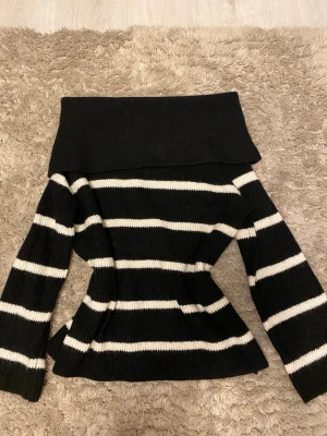 Svart off shoulder stickad tröja - Säljer en svart stickad tröja med bred off shoulder-krage och vita horisontella ränder. Tröjan har lång ärm och figurnära passform, perfekt för dig som gillar en trendig och bekväm stil. Materialet känns mjukt och stretchigt.