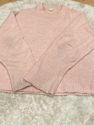 Ljusrosa stickad tröja - Mysig ljusrosa stickad tröja med rund halsringning och breda ribbade muddar vid ärmslut och nederkant. Tröjan har en avslappnad passform och är perfekt för kyliga dagar. Materialet ser mjukt och bekvämt ut, vilket gör den enkel att styla till olika outfits.