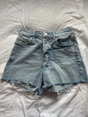 Ljusblå denimshorts med fransig kant - Säljer ett par ljusblå denimshorts med hög midja och rå, fransig kant nertill. Klassisk femficksmodell med knapp och dragkedja framtill. Perfekta för sommaren och ger en avslappnad vibe samtidigt som de är bekväma. Använd endast en gång. Köpa på H&M omedveten om nypris men säljer för 39 kr. 