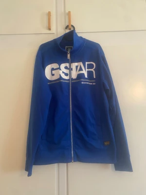 Blå zip hoodie från G-Star RAW - Snygg blå hoodie med dragkedja från G-Star RAW. Stor vit logga på bröstet och liten patch nere vid midjan. Hög krage och långa ärmar. Tillverkad i mjukt material som känns skönt mot huden. Perfekt för en avslappnad streetwear-look.