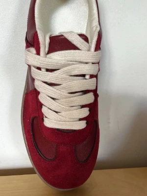 Vinröda sneakers med mockadetaljer - Snygga sneakers i vinrött med ovandel i mocka och textil. Skorna har vita, breda skosnören och en brun gummisula. Stilen är klassisk och passar perfekt till streetwear eller casual outfits.