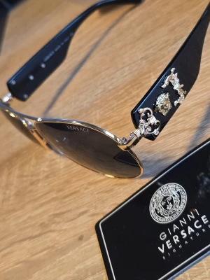 Svarta Versace solglasögon med gulddetaljer - Snygga svarta solglasögon från Versace med guldfärgade detaljer och ikoniska Medusa-emblemet på skalmarna. Glasen är mörka och bågarna har en exklusiv känsla med metallinslag. Perfekta för dig som vill sticka ut med lyxig accessoar.
