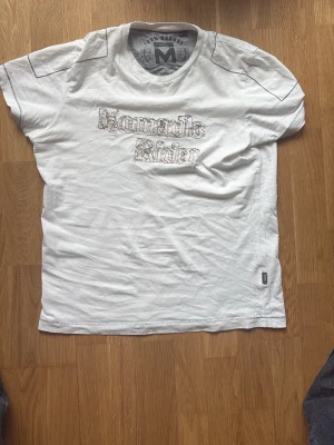 Vit t-shirt av Nomadic rider - Säljer en vit t-shirt från Nomadic ridermed tryck på bröstet i gotisk stil. T-shirten har korta ärmar och en klassisk passform. Perfekt till jeans eller shorts för en avslappnad look.